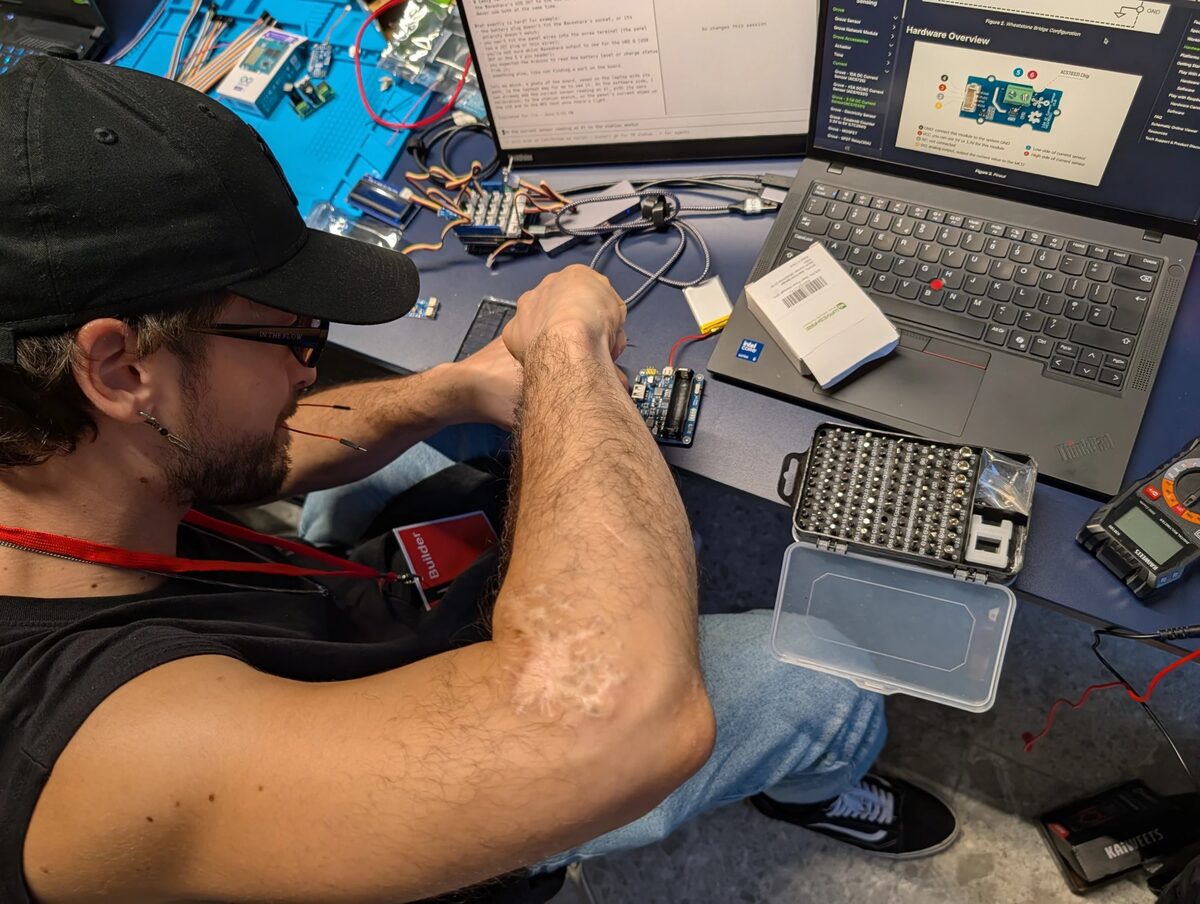 Lorenzo assembling the PoopVolt hardware at hackTurin 2026: wiring a battery charger board next to the Grove modules, with the Grove current sensor documentation open on the laptop, a multimeter and a screwdriver bit set on the desk
