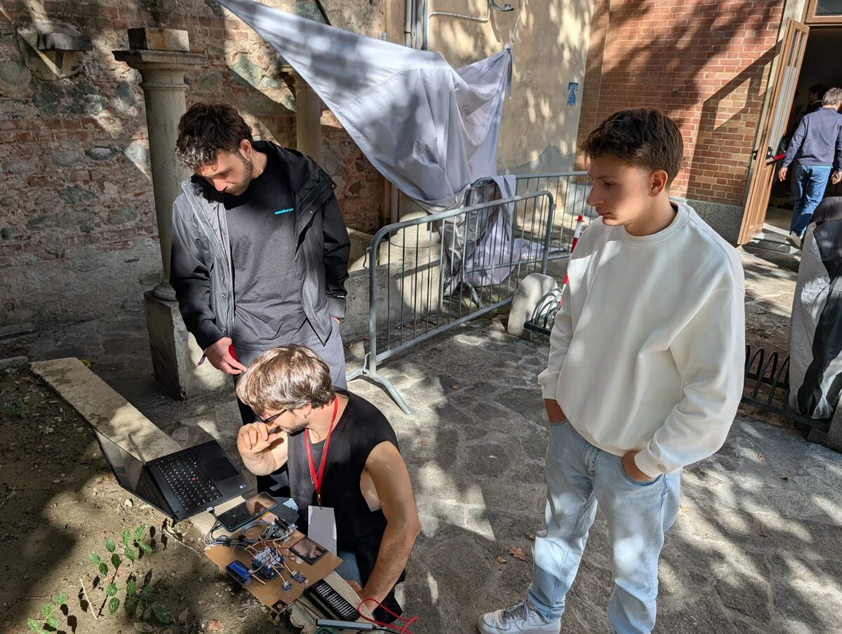 Another debugging session, outdoors in the courtyard: Lorenzo crouching over the prototype board and his laptop, with Giacomo looking on from the left and Federico, in a white sweatshirt, on the right