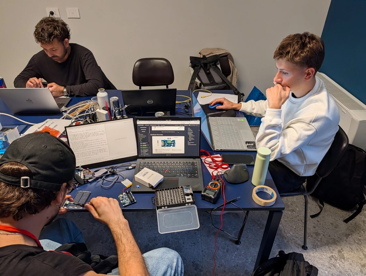The team desk at hackTurin 2026: Giacomo at the top left working on his laptop, Federico on the right in a white sweatshirt looking at his screen, and Lorenzo with his back to the camera wiring the prototype among laptops, a multimeter and a screwdriver bit set