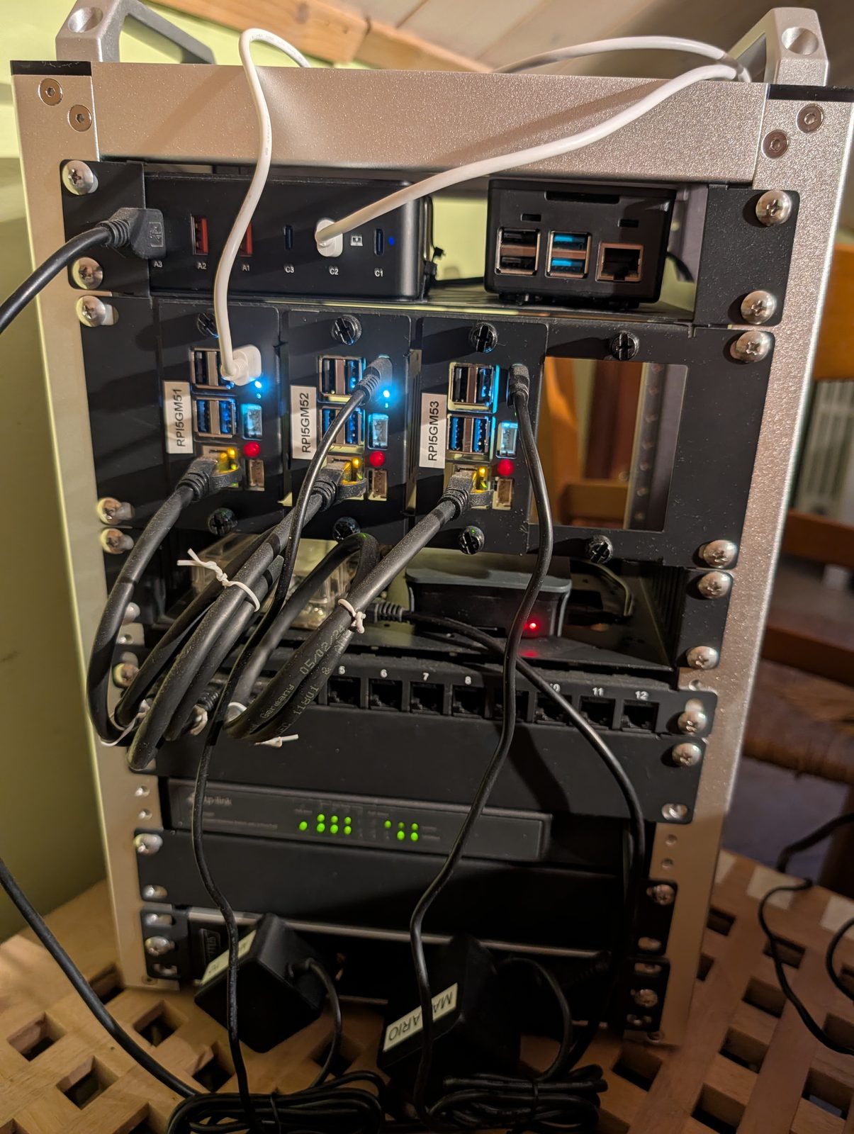 Photo of the k3s-gmhome rack: three Raspberry Pi 5 nodes (rpi5gm51, rpi5gm52, rpi5gm53) in individual carrier trays, cabled down to the PoE+ switch below